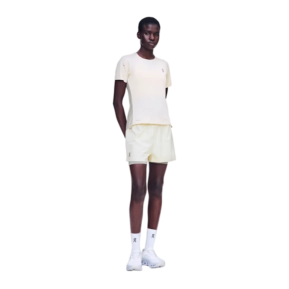 En person står med ansigtet fremad, iført en On Performance-T til kvinder i farvekombinationen Hare/Brume sammen med matchende shorts, hvide sokker og hvide sneakers. Armene er afslappede, og hænderne er placeret bag ryggen, alt sammen på en ensfarvet hvid baggrund.