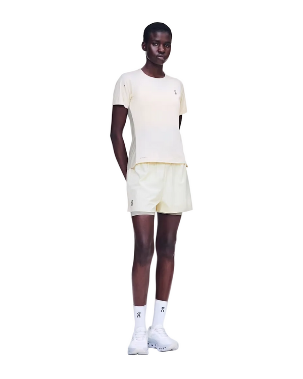 En person står med ansigtet fremad, iført en On Performance-T til kvinder i farvekombinationen Hare/Brume sammen med matchende shorts, hvide sokker og hvide sneakers. Armene er afslappede, og hænderne er placeret bag ryggen, alt sammen på en ensfarvet hvid baggrund.