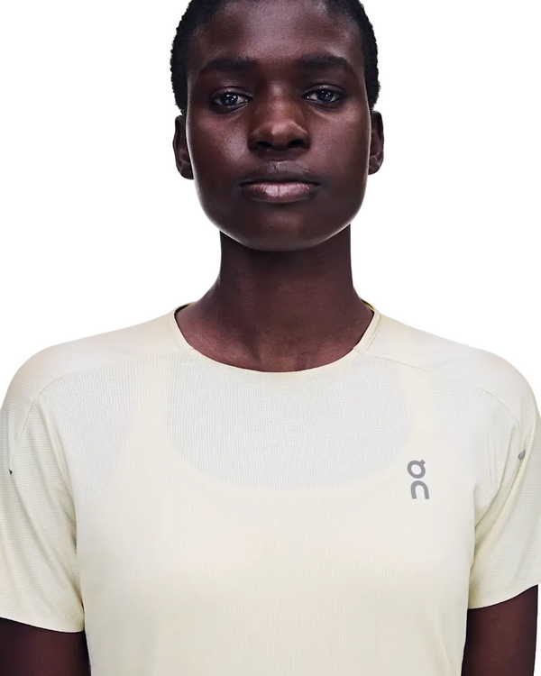 En person med kort hår har en On Performance-T, Dame, Hare/Brume på – en lysbeige, kortærmet sportstrøje med On-logoet på venstre bryst. Denne løbetrøje er fremstillet af svedtransporterende materiale og er ideel til aktive aktiviteter. Personen står foran en ensfarvet hvid baggrund.
