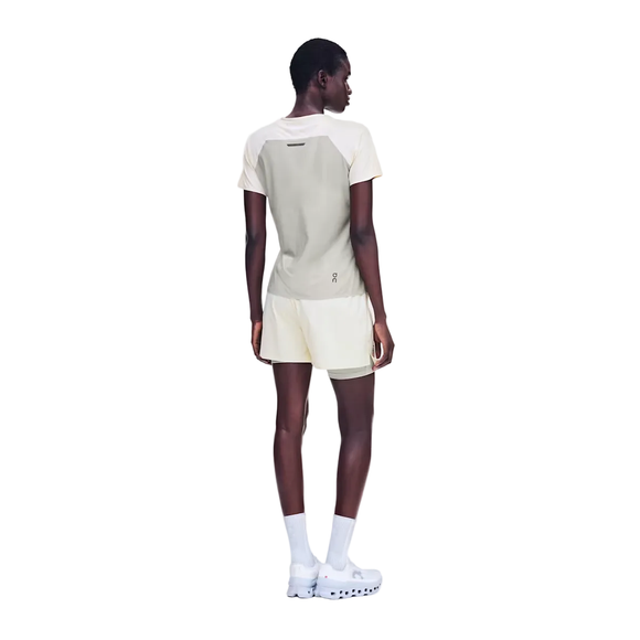 En person med kort hår står med ryggen til kameraet og har en On Performance-T til kvinder i farven Hare/Brume på, der er lavet af svedtransporterende materiale, samt cremefarvede shorts, hvide sokker og hvide sneakers. Baggrunden er ensfarvet hvid.