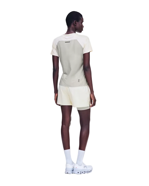 En person med kort hår står med ryggen til kameraet og har en On Performance-T til kvinder i farven Hare/Brume på, der er lavet af svedtransporterende materiale, samt cremefarvede shorts, hvide sokker og hvide sneakers. Baggrunden er ensfarvet hvid.