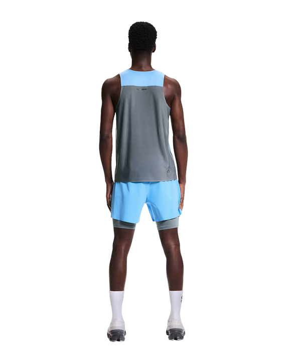En person står med ryggen til kameraet iført On Performance Tank til mænd i Gourami/Rock, lyseblå shorts, hvide sokker og grå sportssko på en almindelig hvid baggrund.