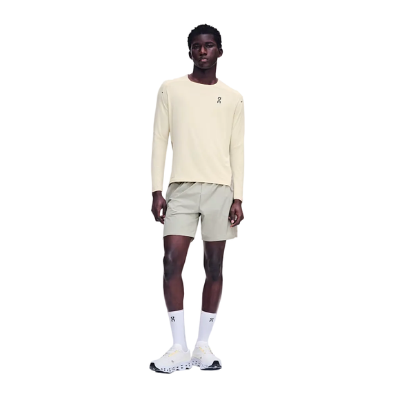 En person står med ansigtet vendt fremad iført en On Performance Long-T, Herre, Hare/Brume – en cremefarvet langærmet løbetrøje af åndbart materiale – kombineret med lyse khaki-shorts, hvide strømper med rund kant og hvide sneakers. Baggrunden er ensfarvet hvid. Der ses On-logoer på trøjen, strømperne og skoene.