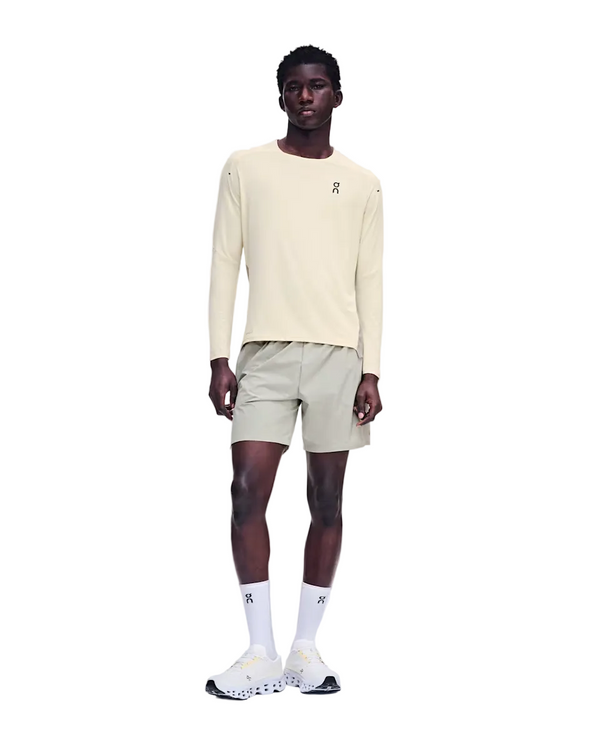En person står med ansigtet vendt fremad iført en On Performance Long-T, Herre, Hare/Brume – en cremefarvet langærmet løbetrøje af åndbart materiale – kombineret med lyse khaki-shorts, hvide strømper med rund kant og hvide sneakers. Baggrunden er ensfarvet hvid. Der ses On-logoer på trøjen, strømperne og skoene.