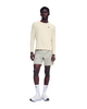 En person står med ansigtet vendt fremad iført en On Performance Long-T, Herre, Hare/Brume – en cremefarvet langærmet løbetrøje af åndbart materiale – kombineret med lyse khaki-shorts, hvide strømper med rund kant og hvide sneakers. Baggrunden er ensfarvet hvid. Der ses On-logoer på trøjen, strømperne og skoene.