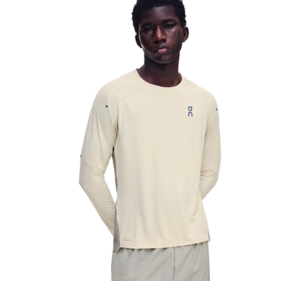 En mand står med ansigtet vendt fremad iført On Performance Long-T til mænd i farven Hare/Brume, der er fremstillet af åndbart materiale. Den lysbeige langærmede trøje har On-logoet på brystet og er kombineret med matchende beige bukser. Han har hænderne bag ryggen og et neutralt udtryk i ansigtet.