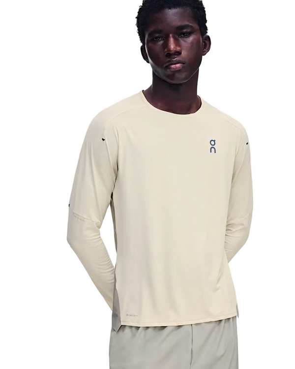 En mand står med ansigtet vendt fremad iført On Performance Long-T til mænd i farven Hare/Brume, der er fremstillet af åndbart materiale. Den lysbeige langærmede trøje har On-logoet på brystet og er kombineret med matchende beige bukser. Han har hænderne bag ryggen og et neutralt udtryk i ansigtet.