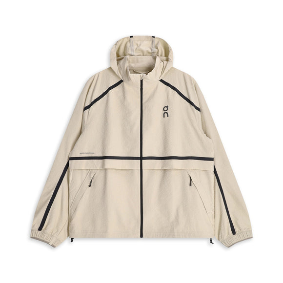 ON Performance Volt Jacket, Herre, Hare er en beige vindjakke med hætte, der vises på en hvid baggrund, designet som en ultralet løbejakke med vindafvisende egenskaber. Den har sorte detaljer ved lynlås og sømme, to forlommer med lynlås, elastiske manchetter, en praktisk hætte, der kan pakkes væk for ekstra bekvemmelighed, og er afsluttet med et lille sort ON-logo på brystet.