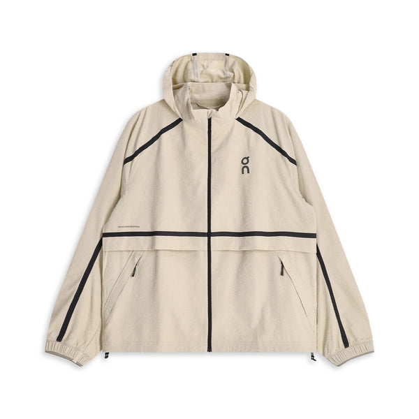 ON Performance Volt Jacket, Herre, Hare er en beige vindjakke med hætte, der vises på en hvid baggrund, designet som en ultralet løbejakke med vindafvisende egenskaber. Den har sorte detaljer ved lynlås og sømme, to forlommer med lynlås, elastiske manchetter, en praktisk hætte, der kan pakkes væk for ekstra bekvemmelighed, og er afsluttet med et lille sort ON-logo på brystet.