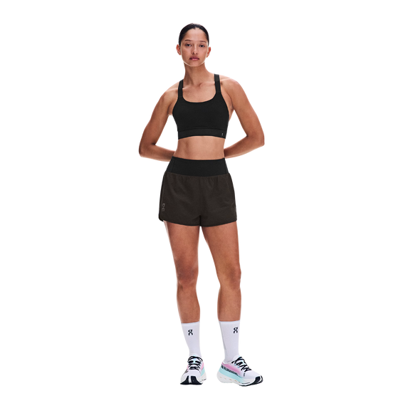 En kvinde står foran en hvid baggrund iført en sort sports-BH, ON Performance Volt Shorts Dame i farven Espresso, hvide strømper med kort skaft og hvide og blågrønne løbesko. Hun har armene over kors og kigger lige frem.