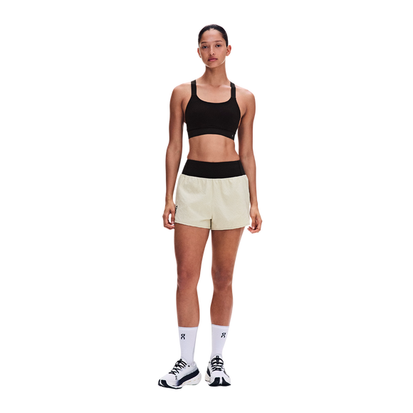 En person står med ansigtet vendt fremad iført en sort sports-BH, ON Performance Volt Shorts Dame Hare med sort linning, hvide strømper og hvide sportssko foran en ensfarvet hvid baggrund.