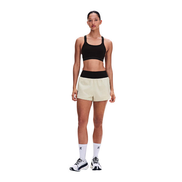 En person står med ansigtet vendt fremad iført en sort sports-BH, ON Performance Volt Shorts Dame Hare med sort linning, hvide strømper og hvide sportssko foran en ensfarvet hvid baggrund.