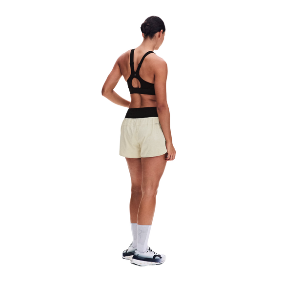 På en ensfarvet hvid baggrund står en kvinde med ryggen til, iført ON’s Performance Volt Shorts Dame Hare med sort linning, en sort sports-BH, hvide sokker og sort-hvide sportssko.