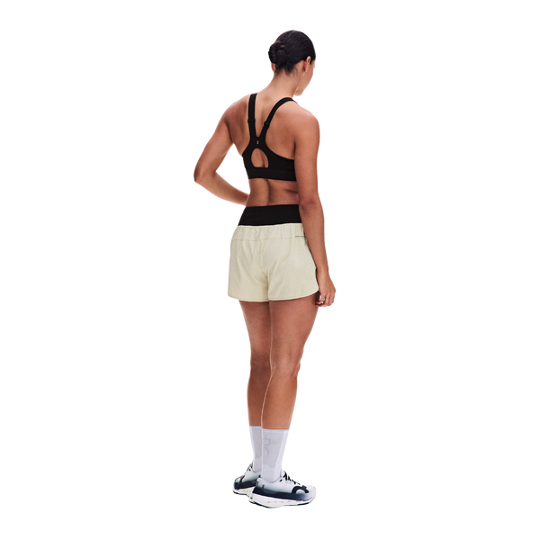 På en ensfarvet hvid baggrund står en kvinde med ryggen til, iført ON’s Performance Volt Shorts Dame Hare med sort linning, en sort sports-BH, hvide sokker og sort-hvide sportssko.