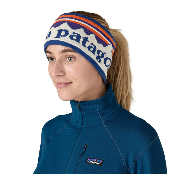 En kvinde iført en blå Patagonia-trøje med lynlås i halsen og et Patagonia Powder Town-pandebånd (unisex) i farven »Hut Tripper Stripe: Birch White«, der er prydet med bjerginspirerede motiver og mærkets navn, kigger let til siden.