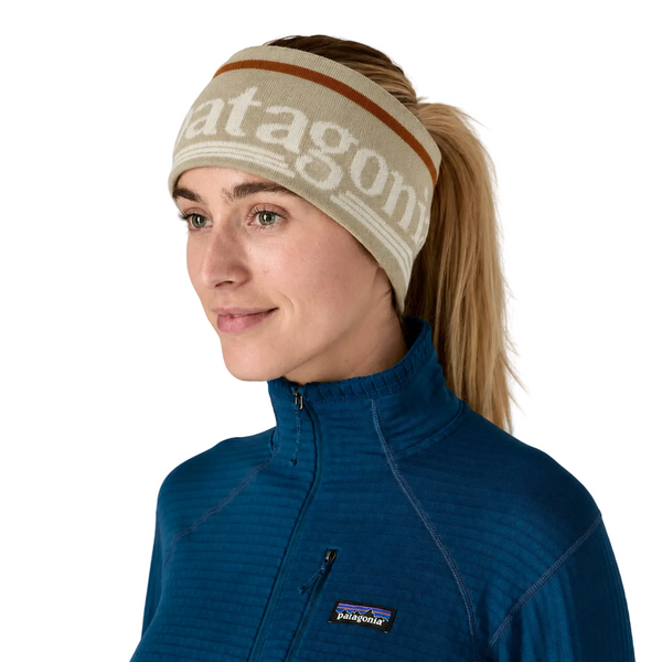 Personen bærer et Patagonia Powder Town-pandebånd (unisex, Hut Tripper-logo: Weathered Stone) fremstillet af genanvendt polyester samt en blå Patagonia-fleecejakke, og har håret sat op i en hestehale. Den hvide baggrund antyder, at personen gør sig klar til vinterløb.