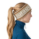 En person med langt blondt hår i en hestehale bærer et Patagonia Powder Town-pandebånd, unisex, med Hut Tripper-logoet i farven »Weathered Stone«, fremstillet af genbrugspolyester, sammen med en blå trøje med lynlås, vist i profil vendt mod højre på en hvid baggrund – perfekt klædt på til vinterløb.