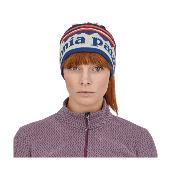 En kvinde med rødt hår, iført en mønstret lilla jakke med lynlås og et Patagonia Powder Town-pandebånd (unisex, Hut Tripper Stripe: Birch White), poserer foran en ensfarvet hvid baggrund.