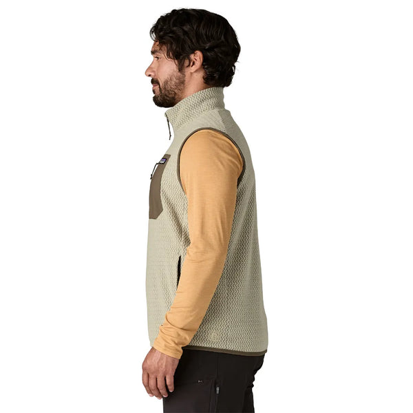 En mand med mørkt hår og skæg, iført en Patagonia R1 Air Vest, Herre, Pelican – en let og åndbar fleecevest til mænd – i beige over en lysebrun langærmet skjorte og mørke bukser, står i profil med ansigtet vendt mod venstre foran en hvid baggrund.
