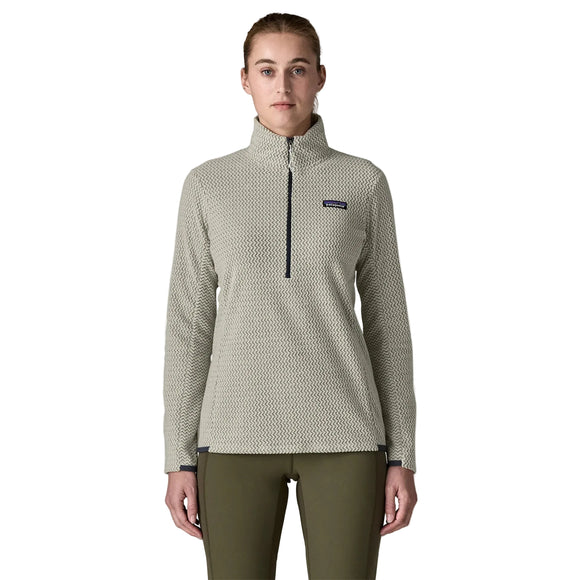 En person står med ansigtet vendt fremad iført en struktureret Patagonia R1 Air Zip Neck-fleece-trøje til kvinder i farven Birch White og olivengrønne bukser foran en ensfarvet lys baggrund. Fleece-trøjen med lynlås i halsen har et lille rektangulært logo på venstre bryst.