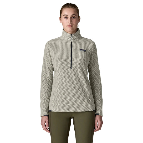 En person står med ansigtet vendt fremad iført en struktureret Patagonia R1 Air Zip Neck-fleece-trøje til kvinder i farven Birch White og olivengrønne bukser foran en ensfarvet lys baggrund. Fleece-trøjen med lynlås i halsen har et lille rektangulært logo på venstre bryst.