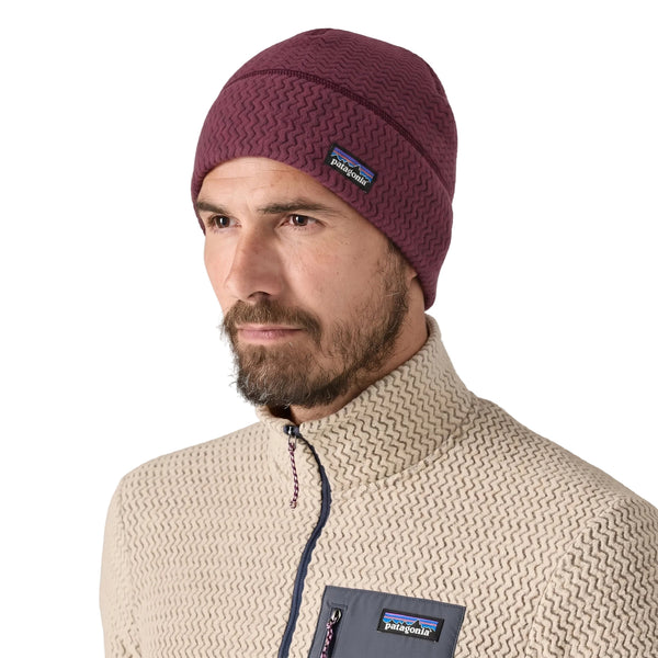 En mand med et velplejet skæg har en Patagonia R1 Air Beanie i farven Berry Fig (unisex) på samt en lysbeige jakke med lynlås, hvor Patagonia-logoet er synligt. Han står vendt let mod venstre foran en ensfarvet hvid baggrund.