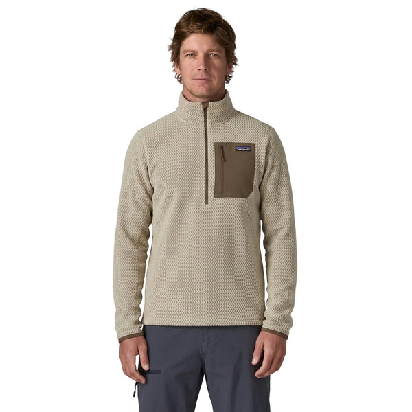 En mand står med ansigtet vendt fremad, iført en Patagonia R1 Air Zip Neck (Herre, Pelican) – en beige pullover med struktur, brystlomme og lynlås – sammen med mørke bukser. Hans hænder hænger ned langs siden. Baggrunden er ensfarvet hvid.