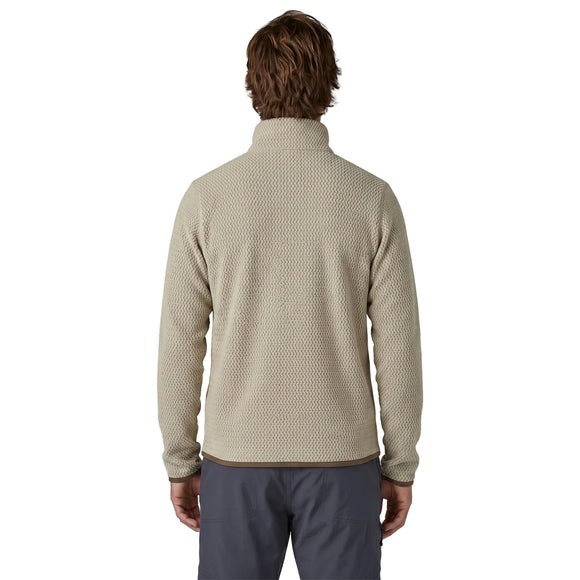 En person med skulderlangt brunt hår, set bagfra, har en Patagonia R1 Air Zip Neck fleece-mellemlagsjakke til mænd med kvartlås i farven Pelican på, der er fremstillet af et struktureret beige stof med brune kanter ved nederkanten og manchetterne. Jakken er kombineret med mørkegrå bukser og fremstår på en hvid baggrund.