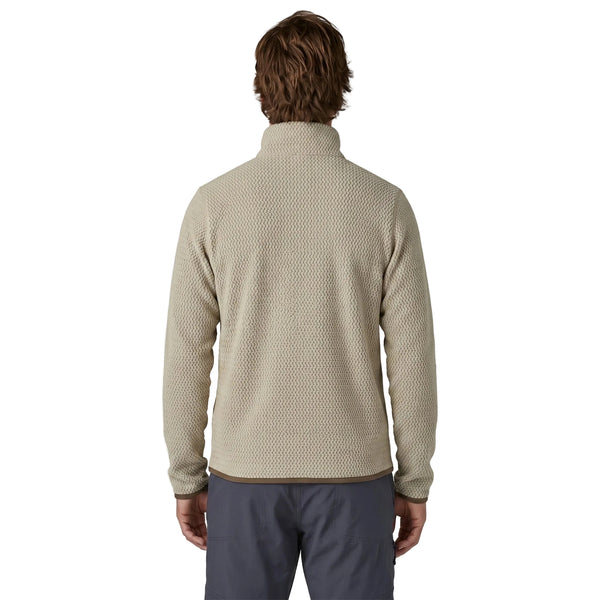 En person med skulderlangt brunt hår, set bagfra, har en Patagonia R1 Air Zip Neck fleece-mellemlagsjakke til mænd med kvartlås i farven Pelican på, der er fremstillet af et struktureret beige stof med brune kanter ved nederkanten og manchetterne. Jakken er kombineret med mørkegrå bukser og fremstår på en hvid baggrund.