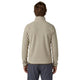 En person med skulderlangt brunt hår, set bagfra, har en Patagonia R1 Air Zip Neck fleece-mellemlagsjakke til mænd med kvartlås i farven Pelican på, der er fremstillet af et struktureret beige stof med brune kanter ved nederkanten og manchetterne. Jakken er kombineret med mørkegrå bukser og fremstår på en hvid baggrund.