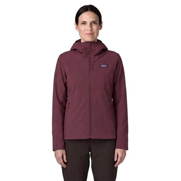 En person står med ansigtet vendt fremad iført en Patagonia R1 CrossStrata-hoodie til kvinder i farven Berry Fig og sorte bukser. Hoodien har en hætte og et lille Patagonia-logo på venstre bryst. Baggrunden er ensfarvet hvid.