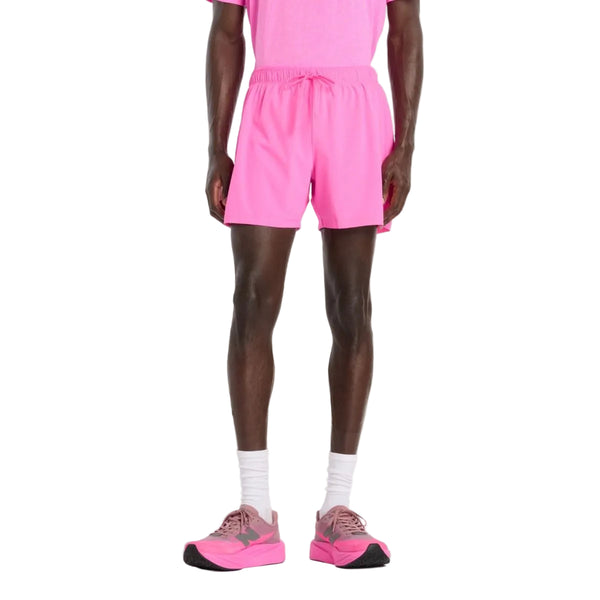 En person står på en hvid baggrund iført lyserøde New Balance RC Short 5" (Herre, Pink Heat), en matchende T-shirt, hvide sokker og lyserøde sneakers. Billedet er beskåret ved skuldrene.