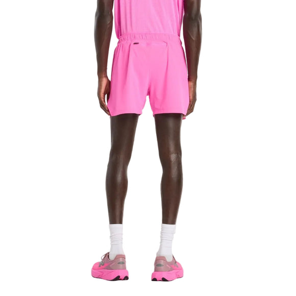 En person står med ryggen til kameraet iført en lyserød skjorte, New Balance RC Short 5" Herre in Pink Heat, hvide sokker og lyserøde sportssko på en almindelig hvid baggrund.