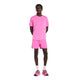 En person står med front mod en almindelig hvid baggrund iført en lyserød T-shirt, New Balance RC Short 5" Herre Pink Heat, hvide sokker og lyserøde sneakers.