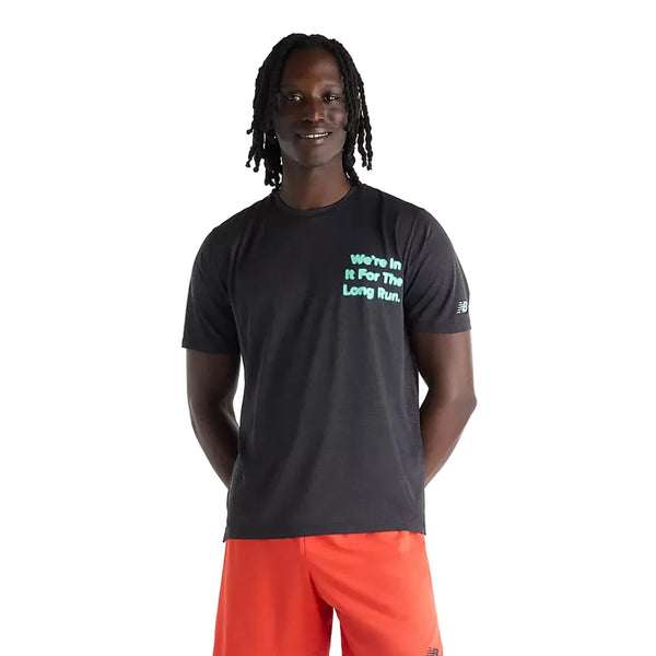 En person står med ansigtet vendt fremad og smiler. Vedkommende har en sort New Balance RC Essential Graphic T-shirt til mænd på med lys mintgrøn tekst og har lys orange shorts på. Hænderne er bag ryggen, og billedet er taget foran en ensfarvet hvid baggrund.