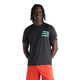 En person står med ansigtet vendt fremad og smiler. Vedkommende har en sort New Balance RC Essential Graphic T-shirt til mænd på med lys mintgrøn tekst og har lys orange shorts på. Hænderne er bag ryggen, og billedet er taget foran en ensfarvet hvid baggrund.