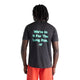 En person med mellemlange dreadlocks står med ryggen til kameraet og har en sort New Balance RC Essential Graphic T-shirt til mænd i farvekombinationen sort/lys mint på, med turkis tekst, hvor der står »We’re in it for the long run«, kombineret med lysorange shorts, foran en ensfarvet hvid baggrund.