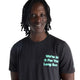 En person med mellemlangt, krøllet hår har en sort New Balance RC Essential Graphic T-shirt til mænd på, hvor der på forsiden står »Were In It For The Long Run.« med lys mintgrøn skrift. Baggrunden er ensfarvet hvid.