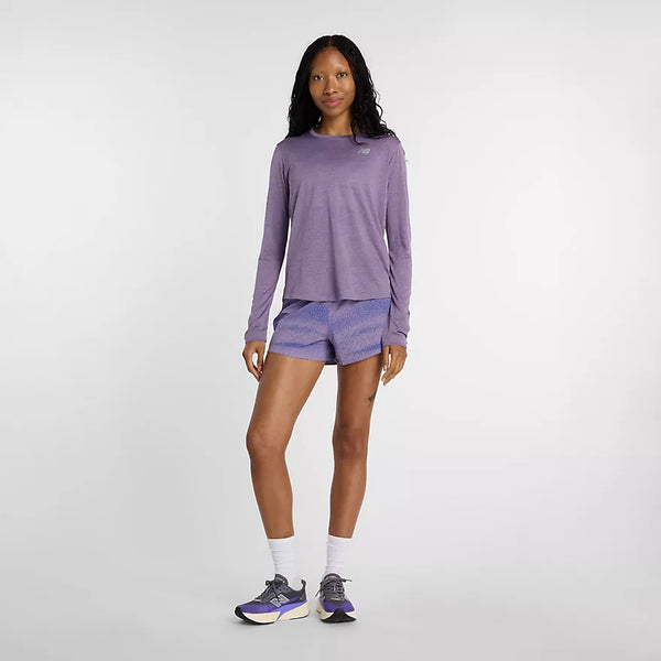 En kvinde står på en ensfarvet baggrund iført New Balance Athletics Long Sleeve til kvinder i Candied Violet Heather, sammen med mønstrede lilla shorts, hvide sokker og New Balance-atletiksko. Hun kigger ind i kameraet med et neutralt udtryk.