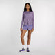En kvinde står på en ensfarvet baggrund iført New Balance Athletics Long Sleeve til kvinder i Candied Violet Heather, sammen med mønstrede lilla shorts, hvide sokker og New Balance-atletiksko. Hun kigger ind i kameraet med et neutralt udtryk.