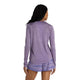 En person med langt krøllet hår, set bagfra mod en hvid baggrund, er iført New Balance Athletics Long Sleeve Dame top i Candied Violet Heather og lyslilla mønstrede shorts.