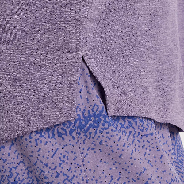 Nærbillede af kanten på New Balance Athletics Long Sleeve, Dame, Candied Violet Heather, med en lille slids i siden, båret over en lilla nederdel med et blåt abstrakt prik- og stregmønster.