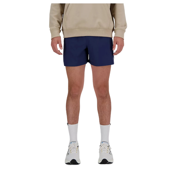 En person står med ansigtet fremad iført en beige sweatshirt, New Balance RC Short 5" Herre Navy/Salte Grey, hvide strømper og hvide sneakers. Billedet er beskåret over skuldrene.