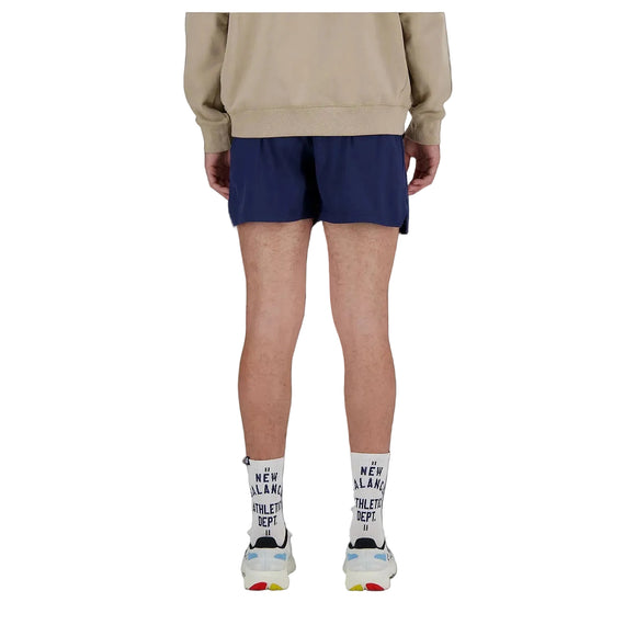 En person står med ryggen til kameraet iført en beige sweatshirt, New Balance RC Short 5" Herre i Navy/Salte Grey, hvide sokker med sort skrift og hvide sneakers med farverige detaljer. Baggrunden er sort.