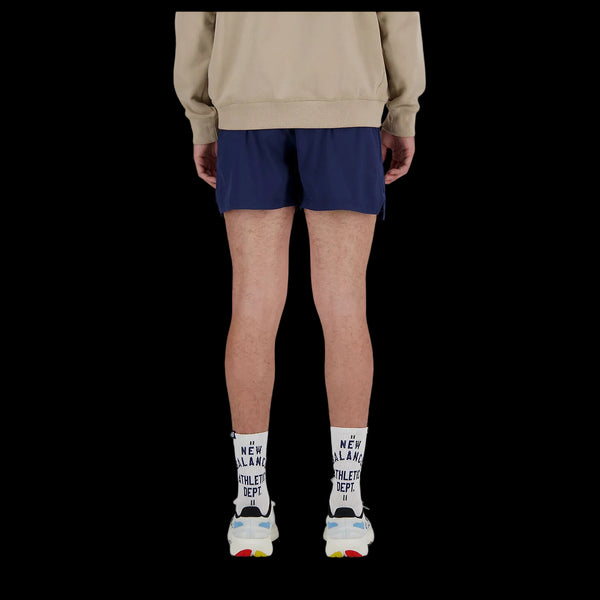En person står med ryggen til kameraet iført en beige sweatshirt, New Balance RC Short 5" Herre i Navy/Salte Grey, hvide sokker med sort skrift og hvide sneakers med farverige detaljer. Baggrunden er sort.