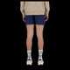 En person står med ryggen til kameraet iført en beige sweatshirt, New Balance RC Short 5" Herre i Navy/Salte Grey, hvide sokker med sort skrift og hvide sneakers med farverige detaljer. Baggrunden er sort.