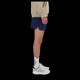 Kun den nederste halvdel af en person i profil er synlig mod en sort baggrund, iført en beige top, hvide tekstsokker, klassiske hvide New Balance-sneakers og RC Short 5" til mænd i Navy/Salte Grey fra New Balance.