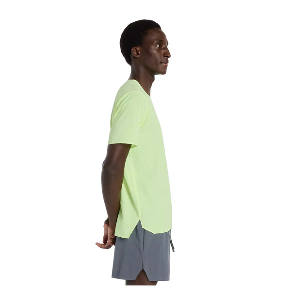 En person i profil vender mod venstre, iført New Balance Race Day Ultra Light T-Shirt til mænd i Afterglow og grå shorts, med armene bag ryggen, på en almindelig hvid baggrund.