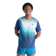 En mand står med hænderne bag ryggen og smiler. Han har en blå New Balance Race Day Ultra Light Printed T-shirt i farvevarianten »Altitude« på og matchende blå shorts, og billedet er taget foran en ensfarvet hvid baggrund.