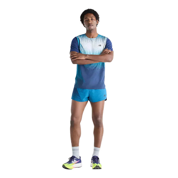En mand står med armene over kors og har en New Balance Race Day Ultra Light Printed T-shirt, Herre, Altitude på – kombineret med matchende blå shorts, grå sokker og neonfarvede løbesko. Baggrunden er helt hvid.